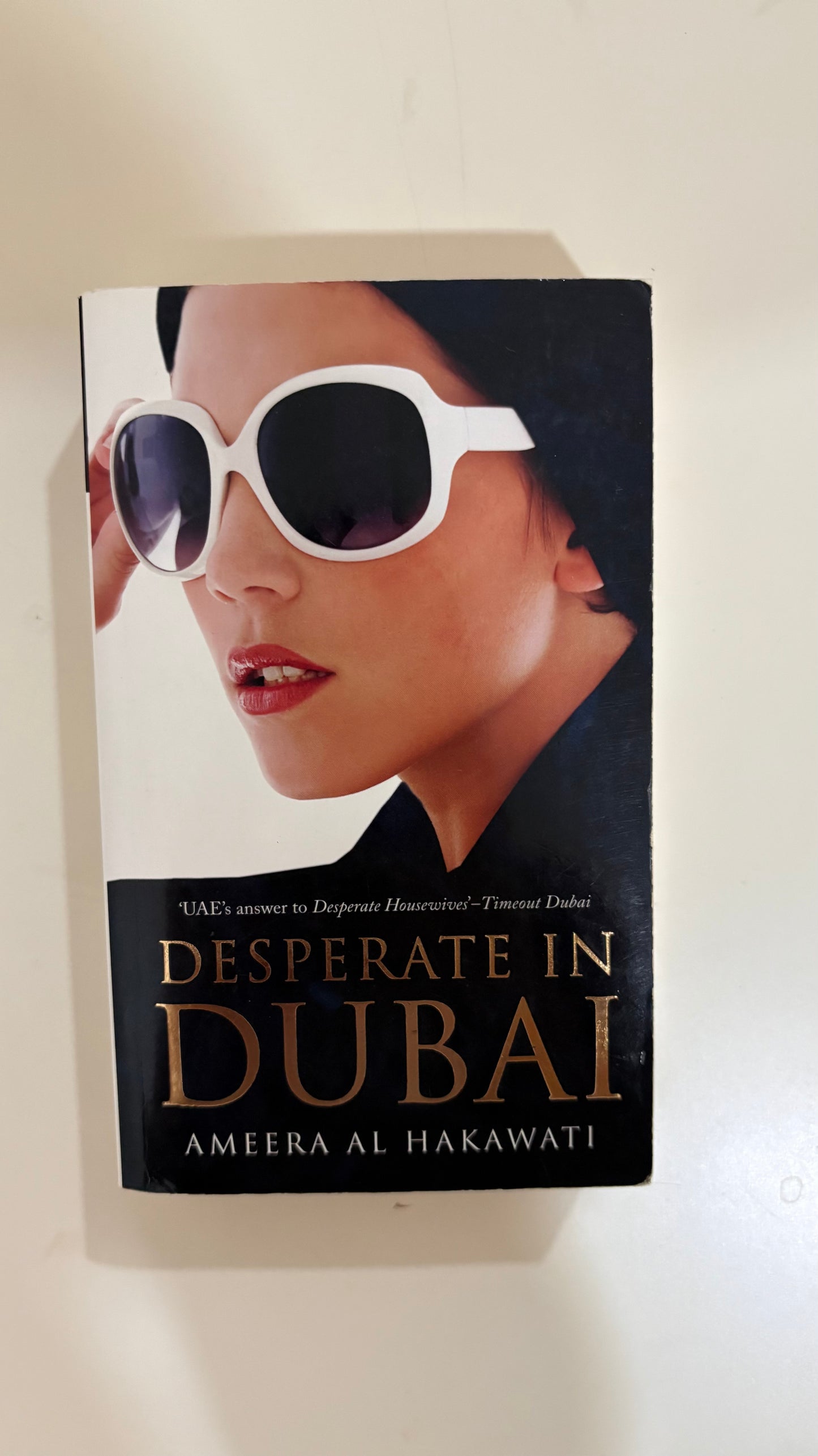 The book cover of 'Desperate in Dubai' by Ameera Al Hakawati, featuring a close-up of a woman wearing large white sunglasses against a black background, with the title in gold lettering.