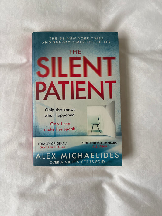 The book cover of 'The Silent Patient' by Alex Michaelides, featuring a blue and white design with bold red title text, photographed on a white fabric background.