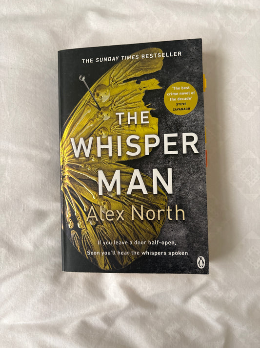 The Whisper Man book by Alex North, featuring a black and yellow cover with a winged insect illustration, photographed on a white textured fabric background.