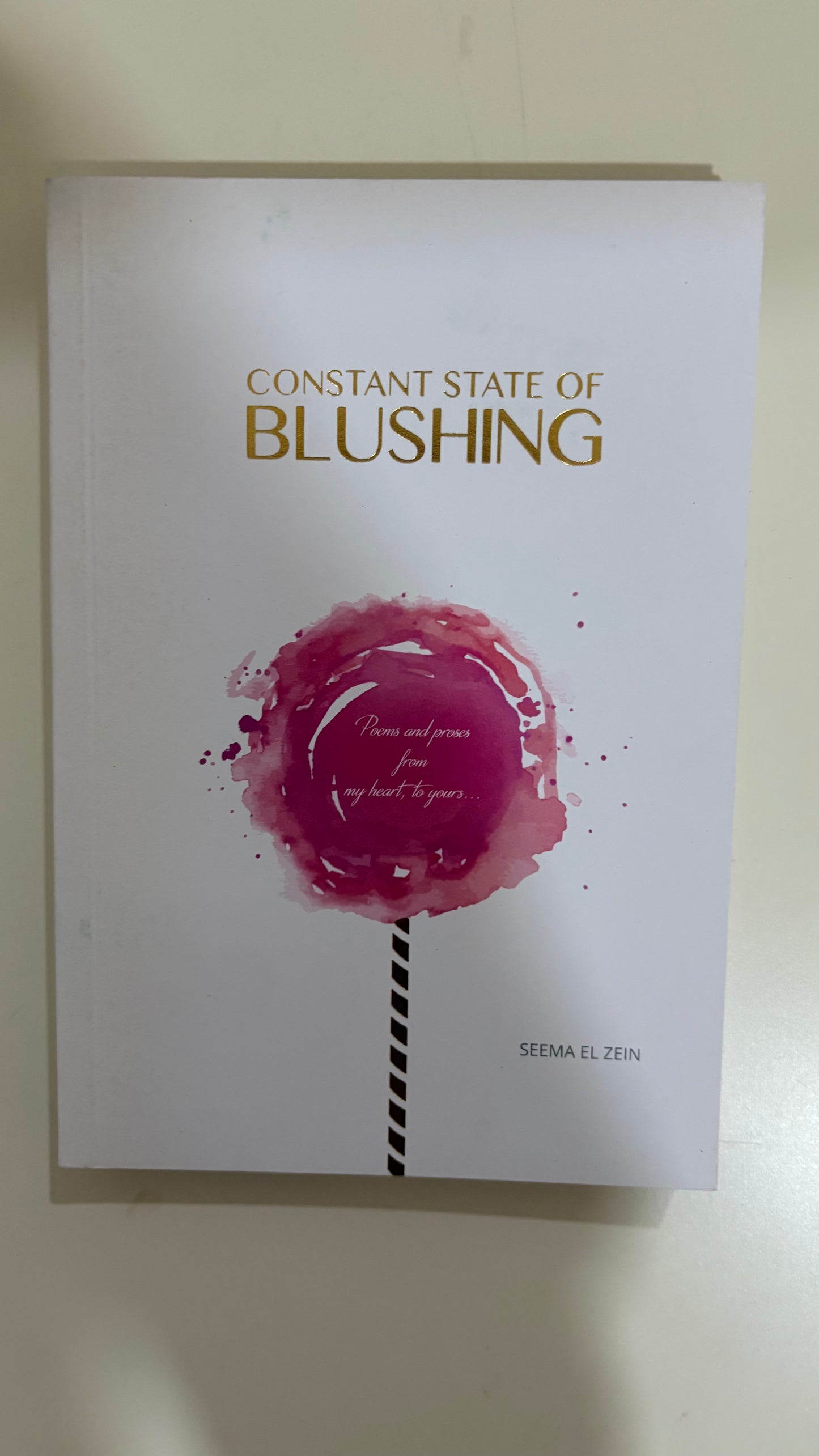 A book titled 'Constant State of Blushing' by Sema Elen, featuring a white cover with a pink watercolor lollipop graphic and gold lettering.