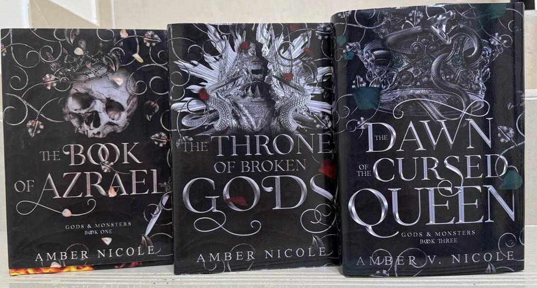 Three books by Amber Nicole titled 'The Book of Azrael', 'The Throne of Broken Gods', and 'Dawn at the Cursed Queen' on a white surface.