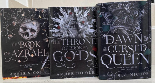 Three books by Amber Nicole titled 'The Book of Azrael', 'The Throne of Broken Gods', and 'Dawn at the Cursed Queen' on a white surface.