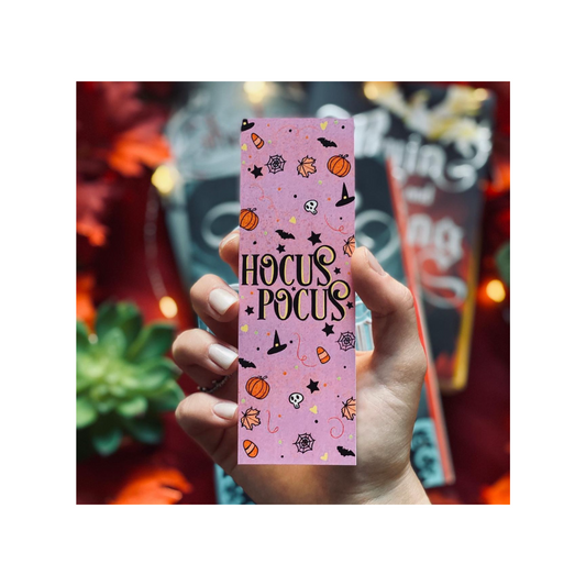 Hand holding a pink 'Hocus Pocus' bookmark with Halloween-themed designs against a festive background.
