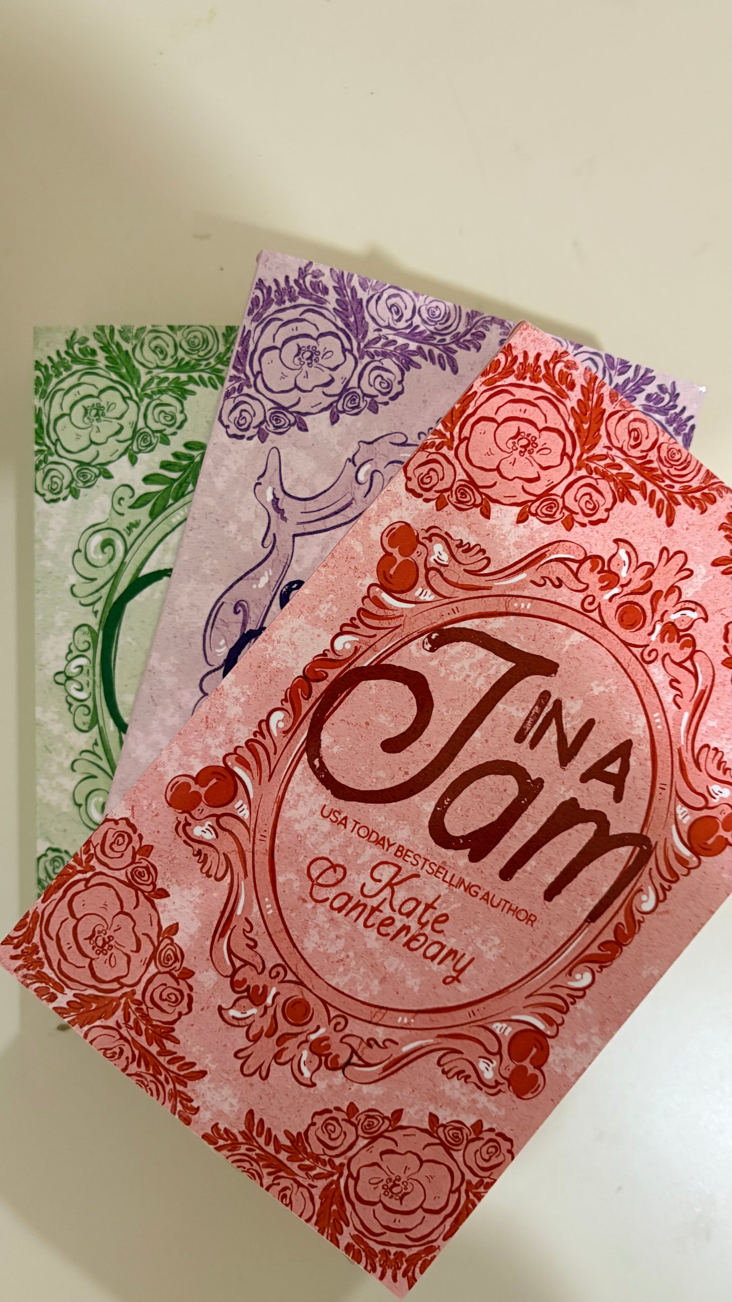 Three decorative book covers fanned out on a light beige surface. The front cover is coral pink with red floral border art and the title 'In a Jam' in large script.