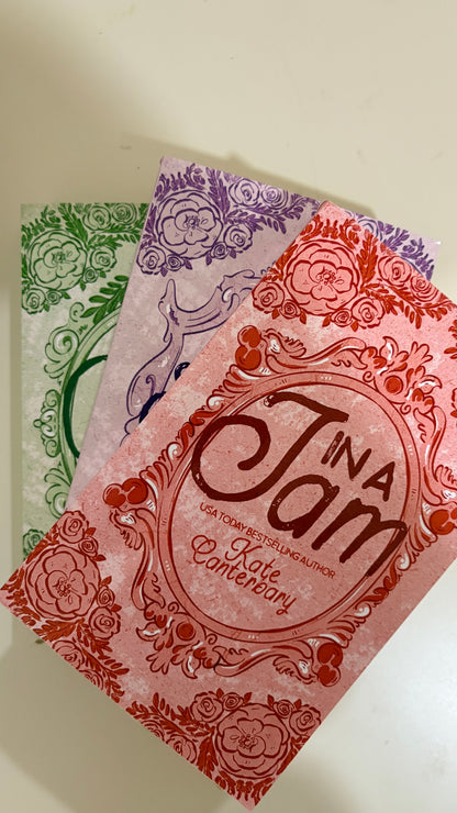Three decorative book covers fanned out on a light beige surface. The front cover is coral pink with red floral border art and the title 'In a Jam' in large script.