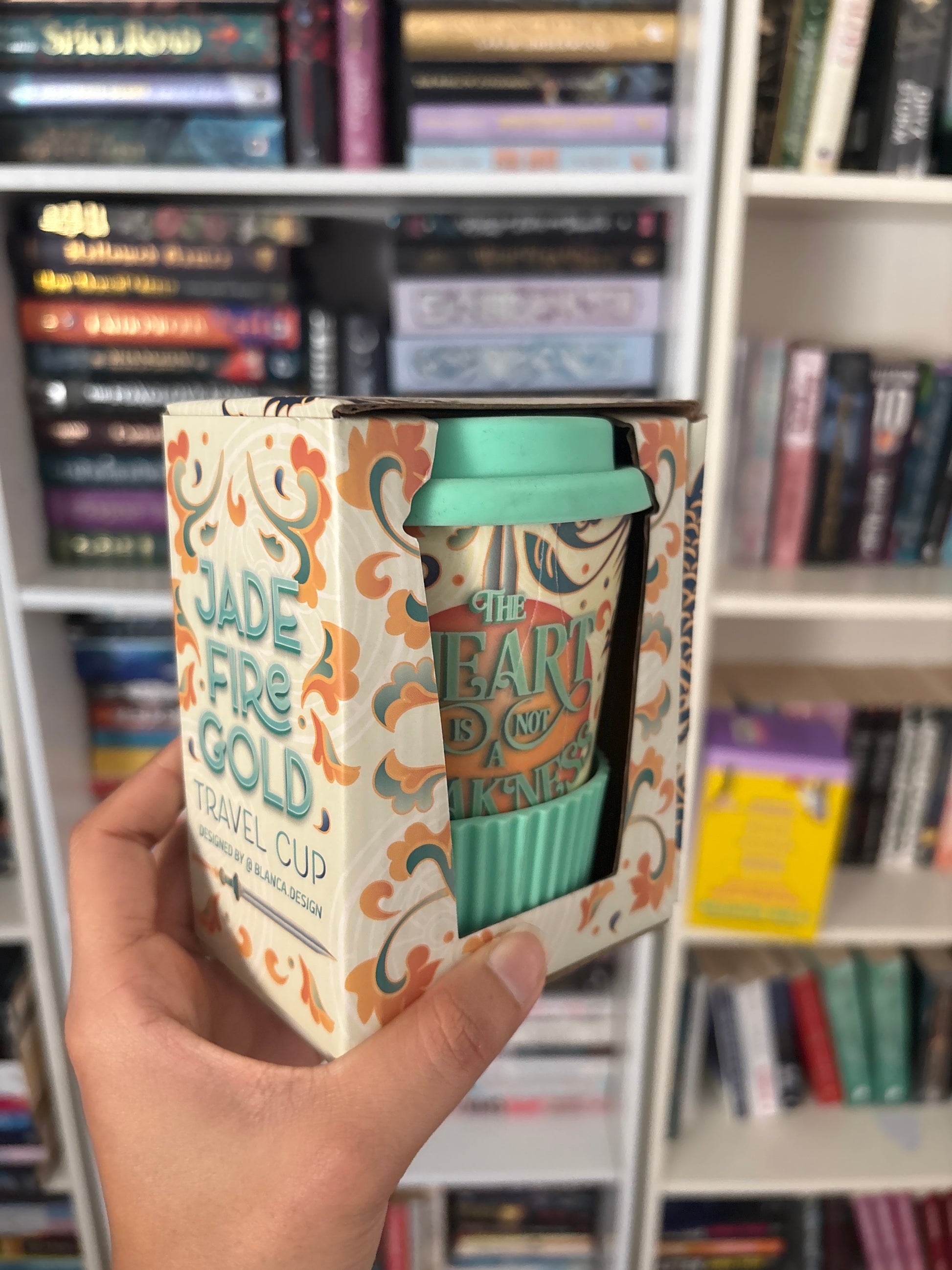 A hand holds a decorative cardboard box containing a mint green travel cup with a matching lid. The box and cup feature a cream background with orange and teal filigree patterns and the text 'Jade Fire Gold' and 'The Earth Is Not Yours.