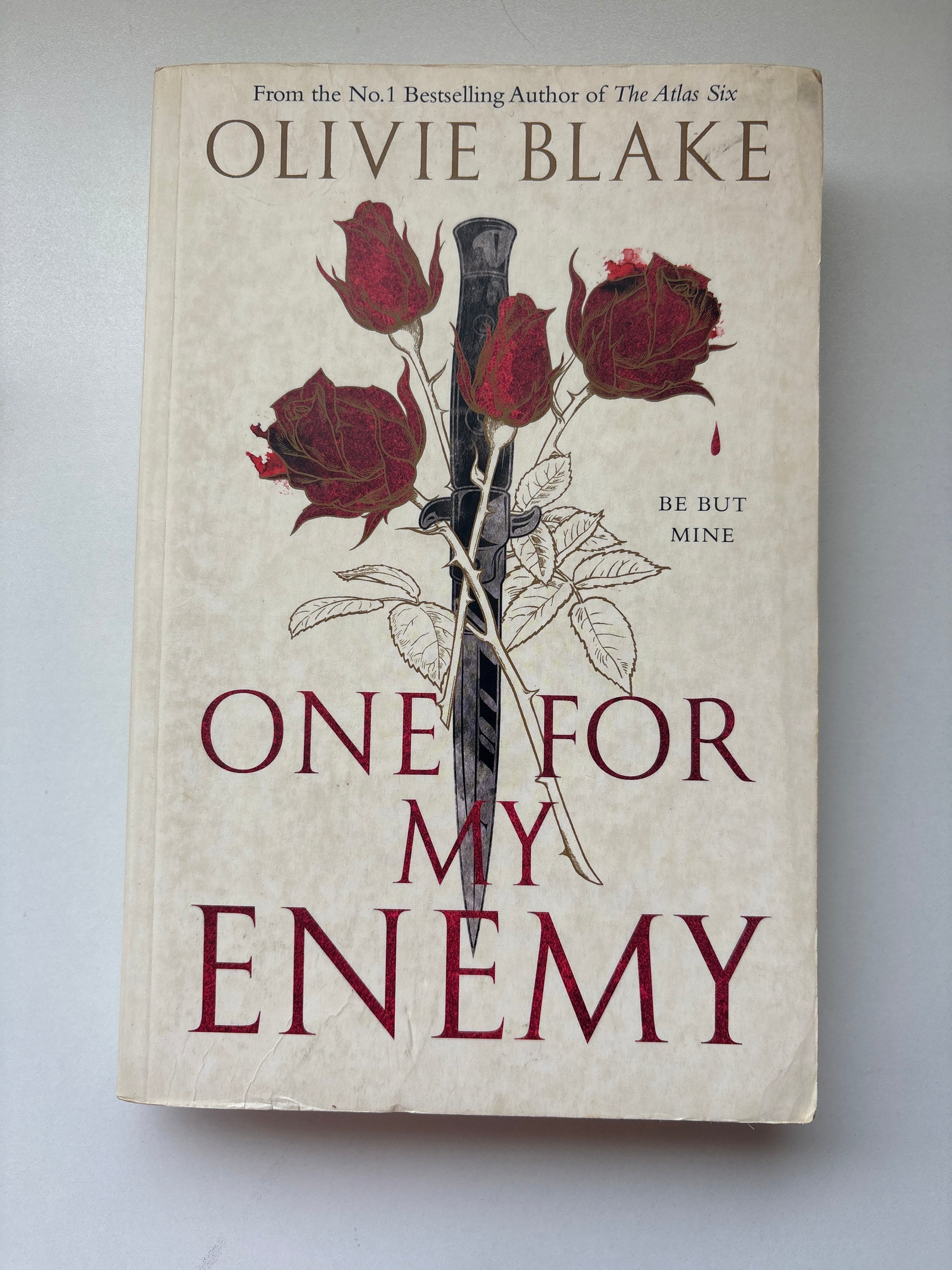 The front cover of the book 'One For My Enemy' by Olivie Blake, featuring a beige background with dark red roses, a black-and-white dagger, and dark red title text.