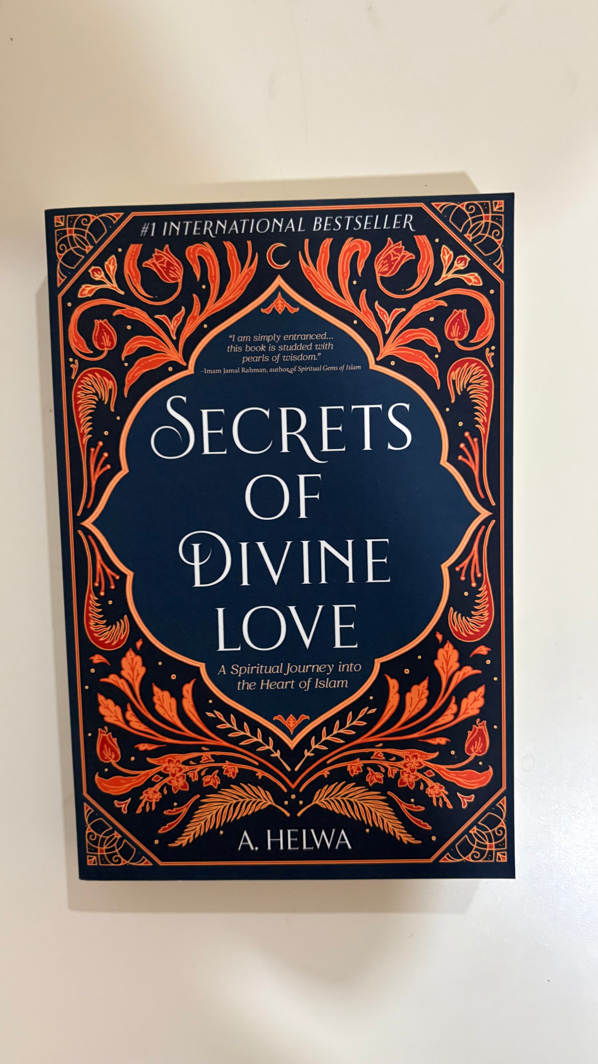 The book cover of 'Secrets of Divine Love' by A. Helwa, featuring a dark navy blue background with intricate orange and gold ornate border designs and white text.