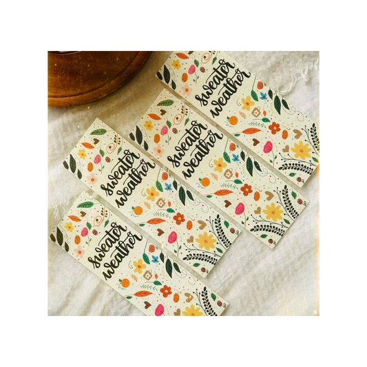 Set of decorative bookmarks with 'sweater weather' text and floral designs on a textured surface.