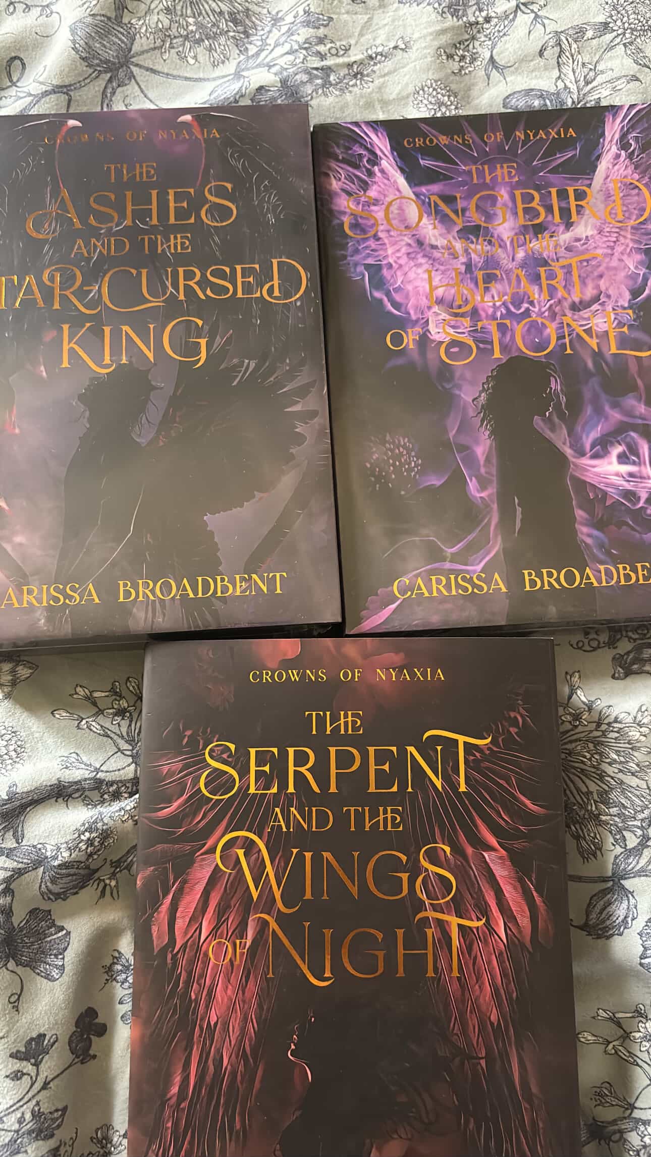Three books titled 'The Ashes and the Star-Cursed King', 'The Songbird and the Heart of Stone', and 'The Serpent and the Wings of Night' by Carissa Broadbent on a patterned fabric background.
