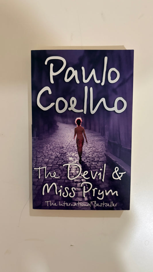 The book cover of 'The Devil & Miss Prym' by Paulo Coelho, featuring a dark purple-toned illustration of a figure walking down a cobblestone path, photographed against a plain off-white background.