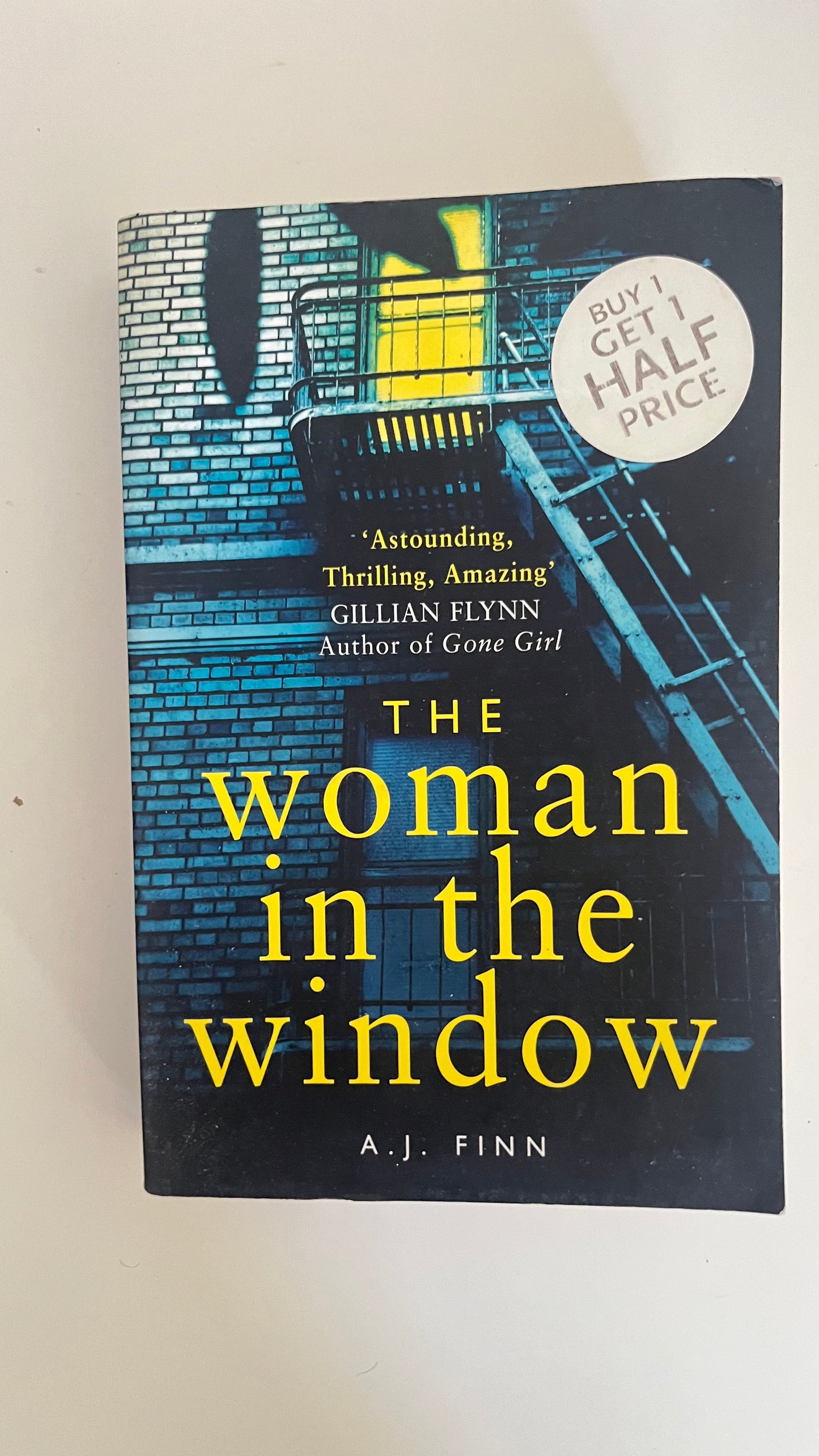 Paperback edition of The Woman in the Window by A.J. Finn with a noir-style cover featuring shadowed blinds and red lettering.

