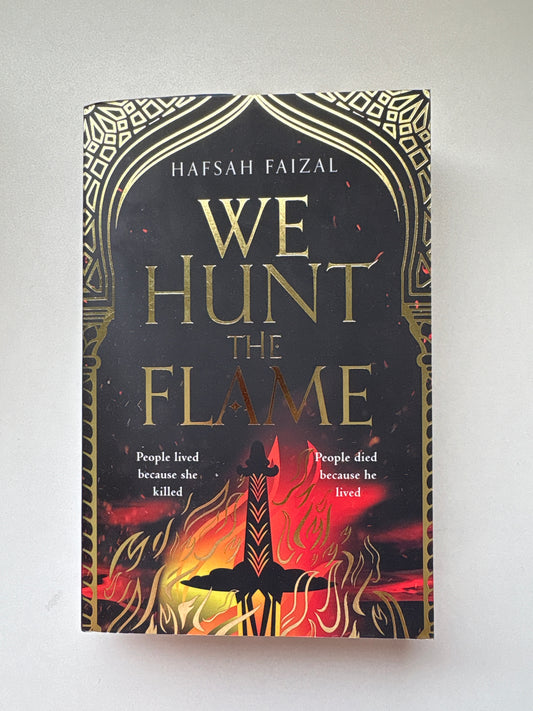 The book cover of 'We Hunt the Flame' by Hafsah Faizal, featuring a black background with gold ornate borders, gold lettering, and a fiery red and orange illustration on the right side.