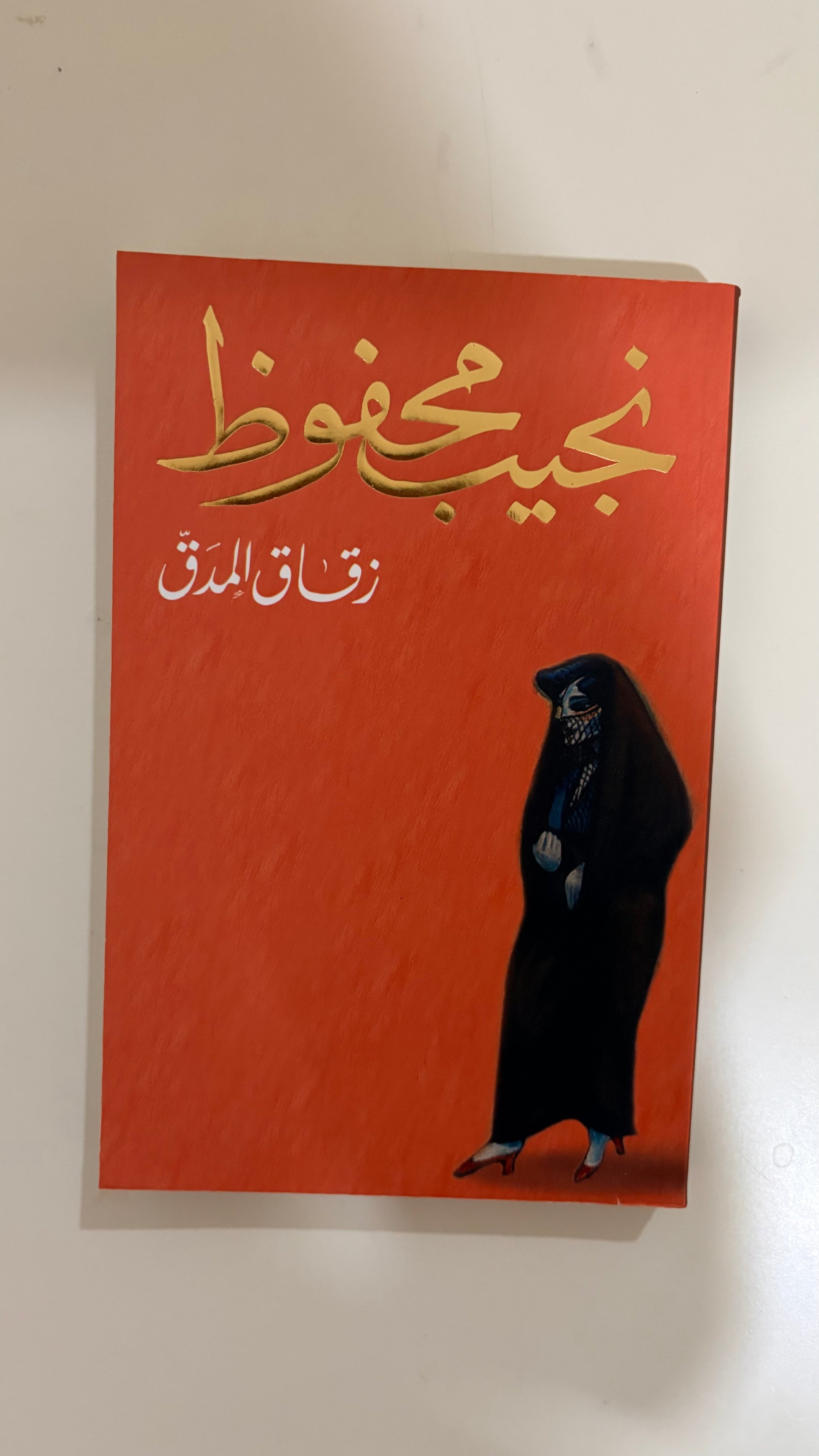 An Arabic book cover with a textured orange-red background, gold Arabic calligraphy at the top, white Arabic text below, and a black-clad figure standing at the lower right. The book is photographed against a plain off-white wall.