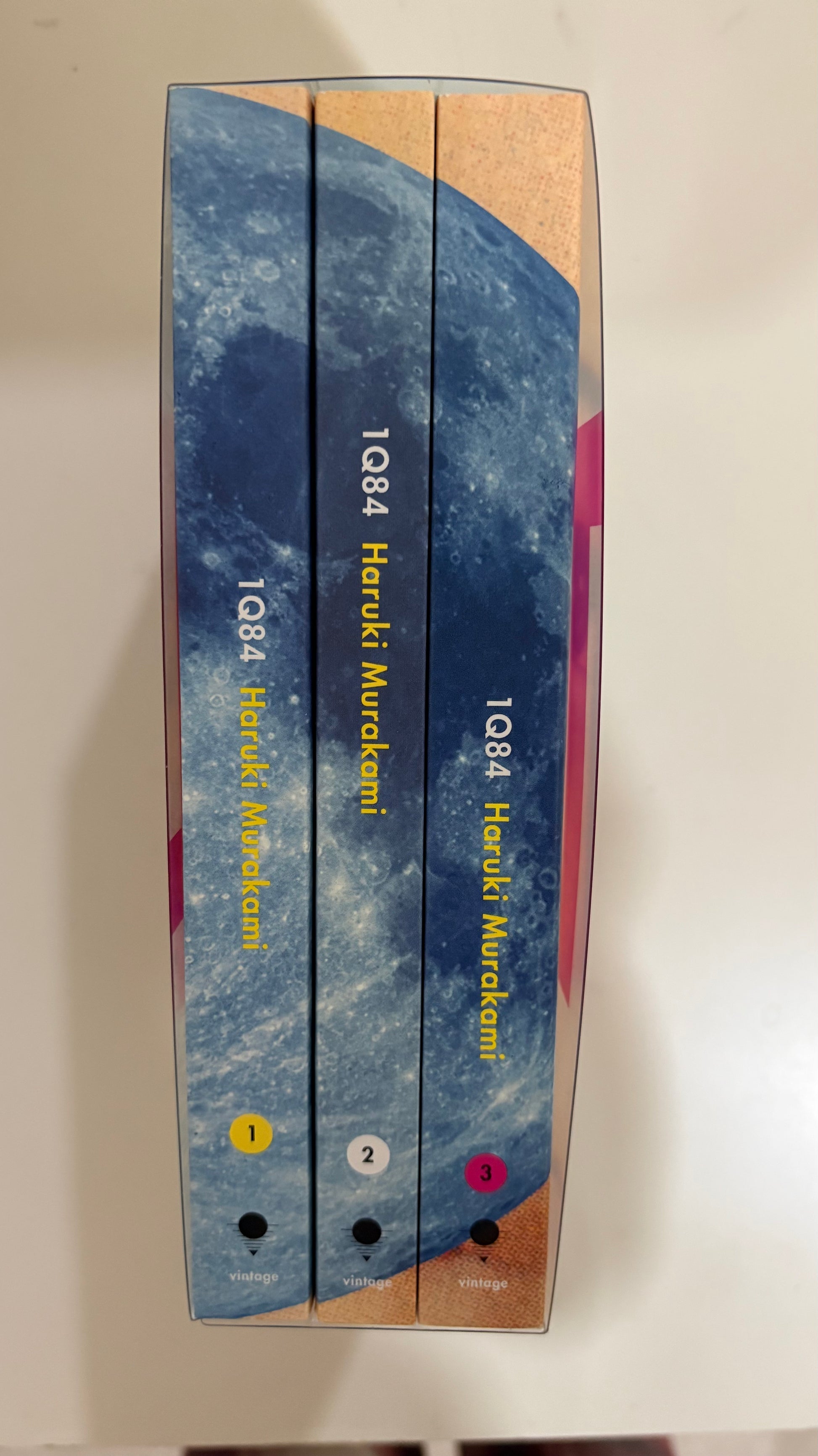 Three books in a hardcover box set by Haruki Murakami, titled 1Q84, with blue and beige covers, photographed on a light surface.