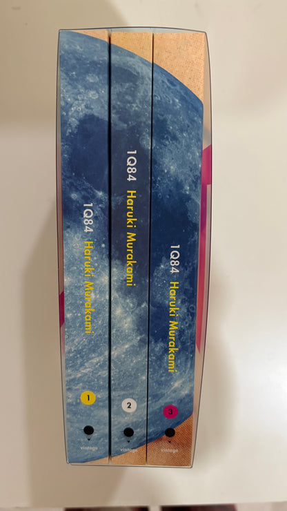 Three books in a hardcover box set by Haruki Murakami, titled 1Q84, with blue and beige covers, photographed on a light surface.